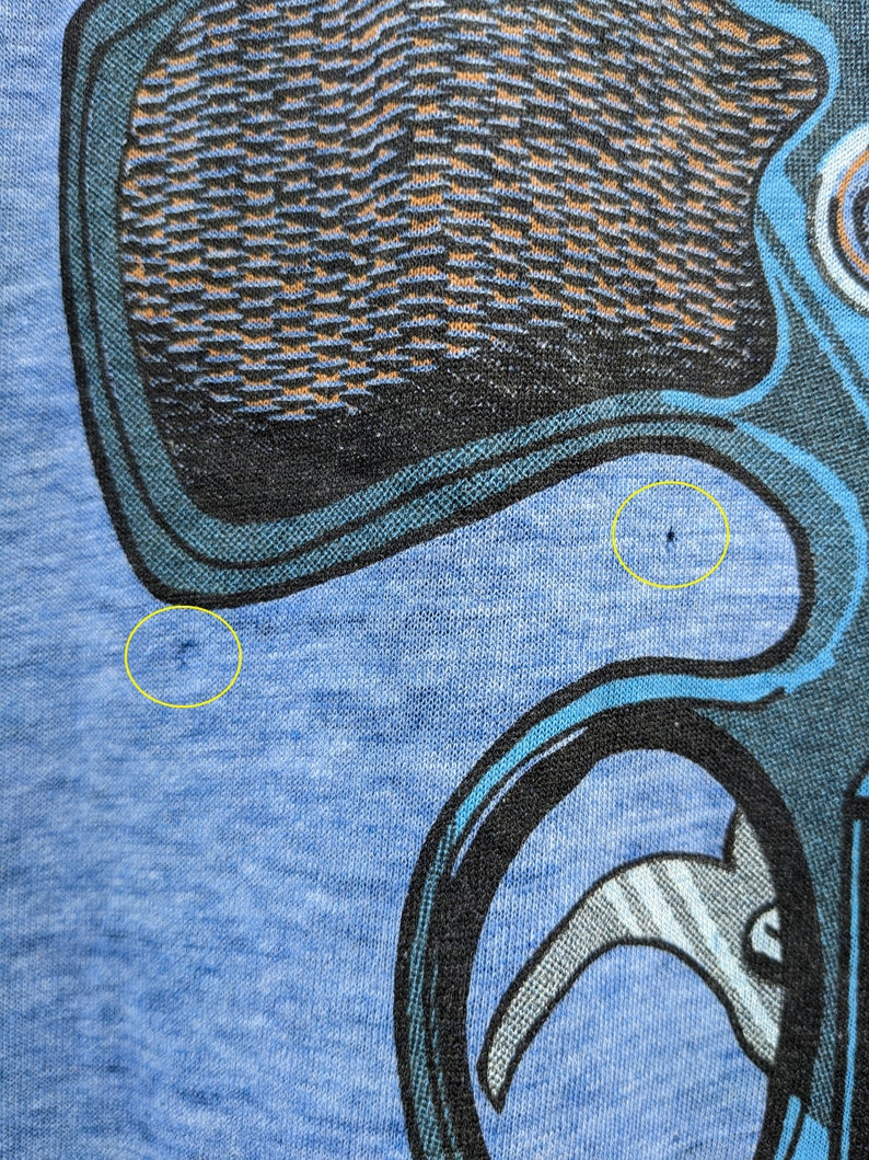 Puede incluir: Una camiseta azul con un estampado gr&aacute;fico negro y azul de un personaje de dibujos animados. El personaje tiene una cabeza grande y un cuerpo peque&ntilde;o. El personaje lleva un sombrero y tiene una nariz grande. La camiseta tiene dos peque&ntilde;os agujeros en la tela.