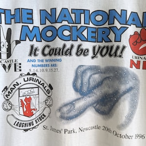 Pu&ograve; includere: Maglietta bianca con una stampa grafica blu e rossa. La grafica presenta il testo "The National Mockery It Could Be You!" con un'illustrazione di una mano e il testo "Newcastle Five" e "Urinal Nil".