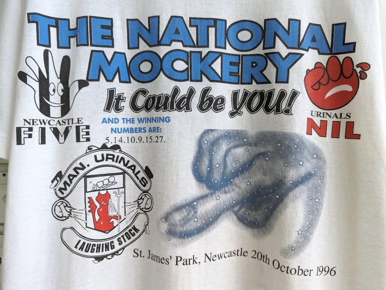 Pu&ograve; includere: Maglietta bianca con una stampa grafica blu e rossa. La grafica presenta il testo "The National Mockery It Could Be You!" con un'illustrazione di una mano e il testo "Newcastle Five" e "Urinal Nil".