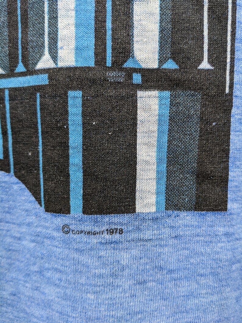 Puede incluir: Una camiseta azul con un dise&ntilde;o geom&eacute;trico en blanco y negro. La camiseta tiene el texto "COPYRIGHT 1978" impreso en la parte inferior.