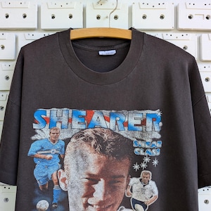 May include: Dark gray t-shirt featuring a graphic of a soccer player with the name "SHEARER" in blue and red lettering. The shirt has a crew neck and short sleeves. The image includes two action shots of the player.