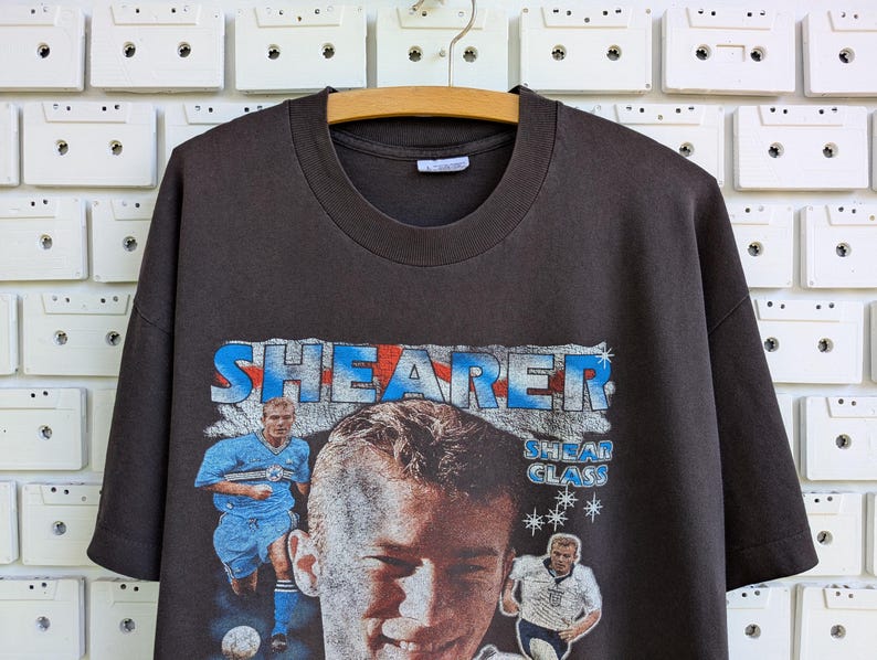 May include: Dark gray t-shirt featuring a graphic of a soccer player with the name "SHEARER" in blue and red lettering. The shirt has a crew neck and short sleeves. The image includes two action shots of the player.
