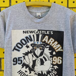 Puede incluir: Camiseta gris con un estampado gr&aacute;fico en blanco y negro de un p&aacute;jaro de dibujos animados con un sombrero con estrellas y rayas. El gr&aacute;fico dice "Newcastle's Toon Army 95-96".