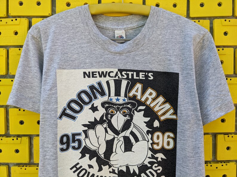 Puede incluir: Camiseta gris con un estampado gr&aacute;fico en blanco y negro de un p&aacute;jaro de dibujos animados con un sombrero con estrellas y rayas. El gr&aacute;fico dice "Newcastle's Toon Army 95-96".