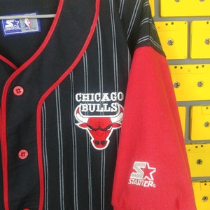 May include: Black and red baseball jersey with white pinstripes. The jersey has a red collar and red trim around the sleeves. The front of the jersey has a red and white Chicago Bulls logo with the text "CHICAGO BULLS". The back of the jersey has the text "STARTER" in white. The jersey is size L.