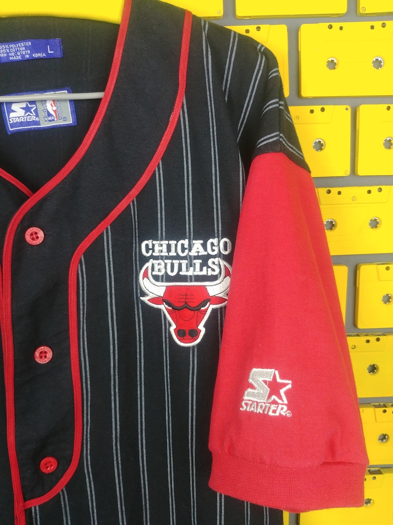May include: Black and red baseball jersey with white pinstripes. The jersey has a red collar and red trim around the sleeves. The front of the jersey has a red and white Chicago Bulls logo with the text "CHICAGO BULLS". The back of the jersey has the text "STARTER" in white. The jersey is size L.