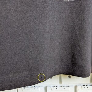 May include: Dark gray t-shirt with the text "THERE IS A GEORDIE" in white and red. The bottom hem of the shirt is visible, with a small hole near the edge. The shirt is made of a soft, textured fabric.