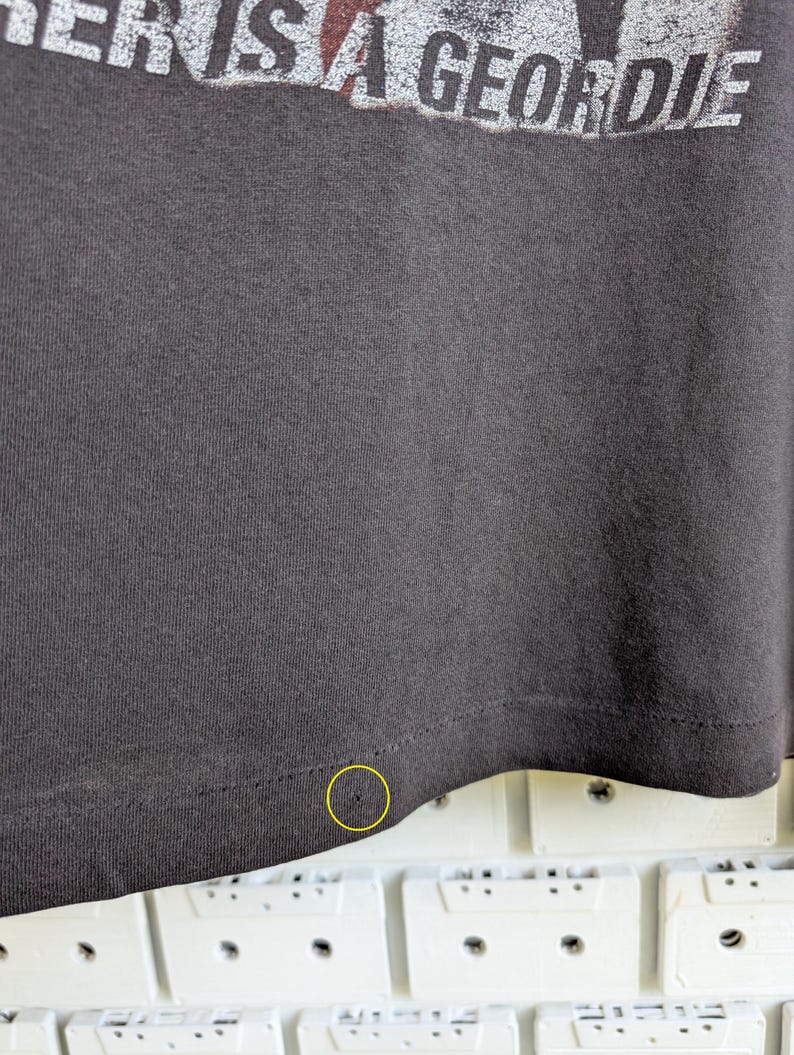 May include: Dark gray t-shirt with the text "THERE IS A GEORDIE" in white and red. The bottom hem of the shirt is visible, with a small hole near the edge. The shirt is made of a soft, textured fabric.