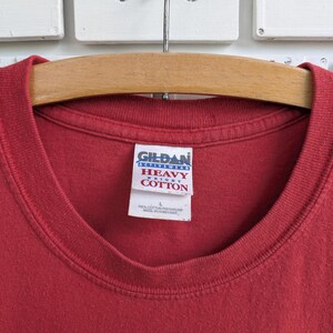 May include: Close-up of a red Gildan Heavy Cotton t-shirt. The shirt is hanging on a wooden hanger. The Gildan tag is visible, showing the brand name and fabric details. The shirt is a solid color.