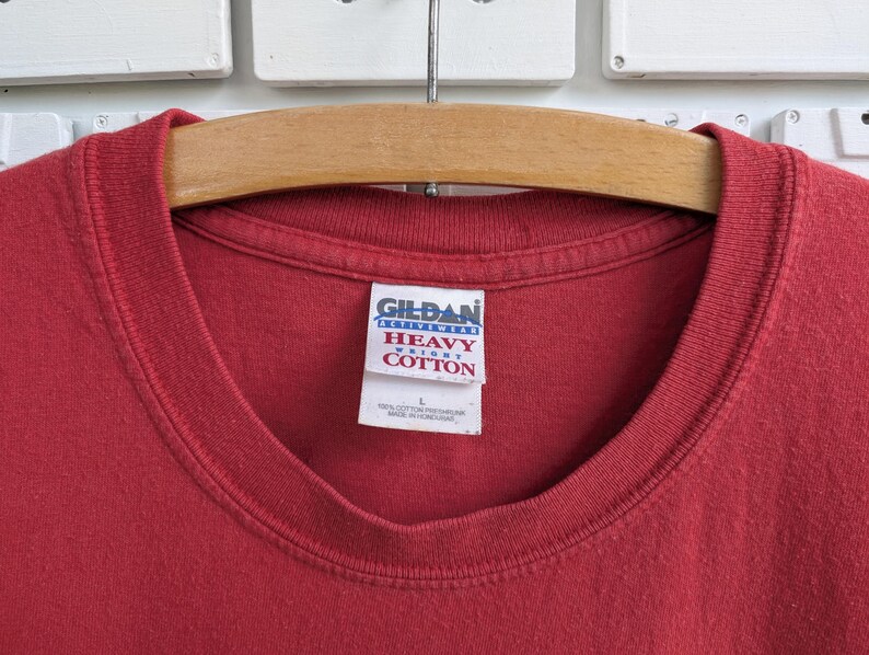 May include: Close-up of a red Gildan Heavy Cotton t-shirt. The shirt is hanging on a wooden hanger. The Gildan tag is visible, showing the brand name and fabric details. The shirt is a solid color.