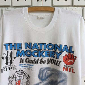 Pu&ograve; includere: Maglietta bianca con una stampa grafica blu e rossa che dice "The National Mockery It Could Be You!" con il testo "Newcastle Five" e "Urinal Nil" in basso.
