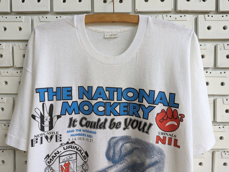 Pu&ograve; includere: Maglietta bianca con una stampa grafica blu e rossa che dice "The National Mockery It Could Be You!" con il testo "Newcastle Five" e "Urinal Nil" in basso.