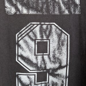 May include: Close-up of a dark gray t-shirt with the word "SHEADER" printed across the top in a distressed, white font. Below is a large number "9" in a white, outlined, geometric design with a star graphic.