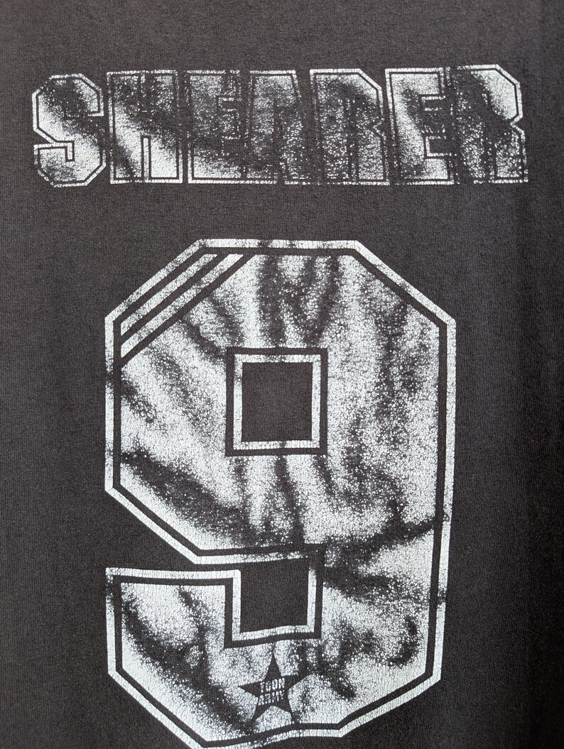 May include: Close-up of a dark gray t-shirt with the word "SHEADER" printed across the top in a distressed, white font. Below is a large number "9" in a white, outlined, geometric design with a star graphic.