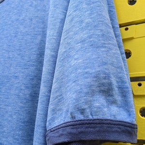 Puede incluir: Una camiseta azul con un ribete acanalado azul marino alrededor del cuello y las mangas. La camiseta tiene un aspecto descolorido y desgastado.
