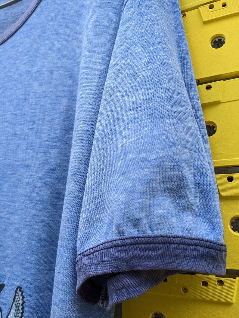 Puede incluir: Una camiseta azul con un ribete acanalado azul marino alrededor del cuello y las mangas. La camiseta tiene un aspecto descolorido y desgastado.