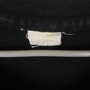 May include: A white tag sewn into a black shirt with the text "ON BACK 100% COTTON MADE IN USA  HOS SIDE"