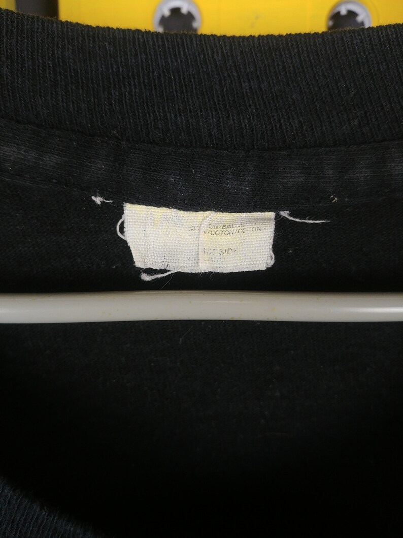 May include: A white tag sewn into a black shirt with the text "ON BACK 100% COTTON MADE IN USA  HOS SIDE"