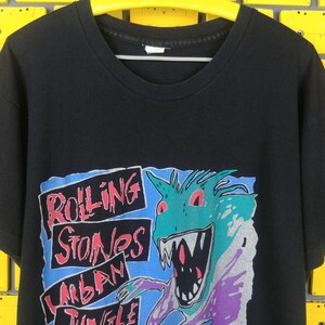May include: Black t-shirt with a graphic print of a cartoon monster with red eyes and a toothy grin. The text "Rolling Stones Urban Jungle" is printed on the shirt.