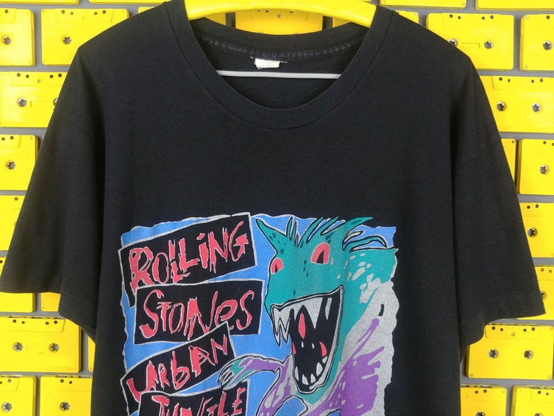 May include: Black t-shirt with a graphic print of a cartoon monster with red eyes and a toothy grin. The text "Rolling Stones Urban Jungle" is printed on the shirt.