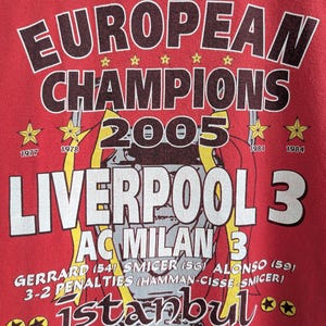 May include: Red t-shirt with a graphic commemorating Liverpool's 2005 Champions League victory. The design includes the words "EUROPEAN CHAMPIONS 2005", team names, scores, and the location and date of the match.