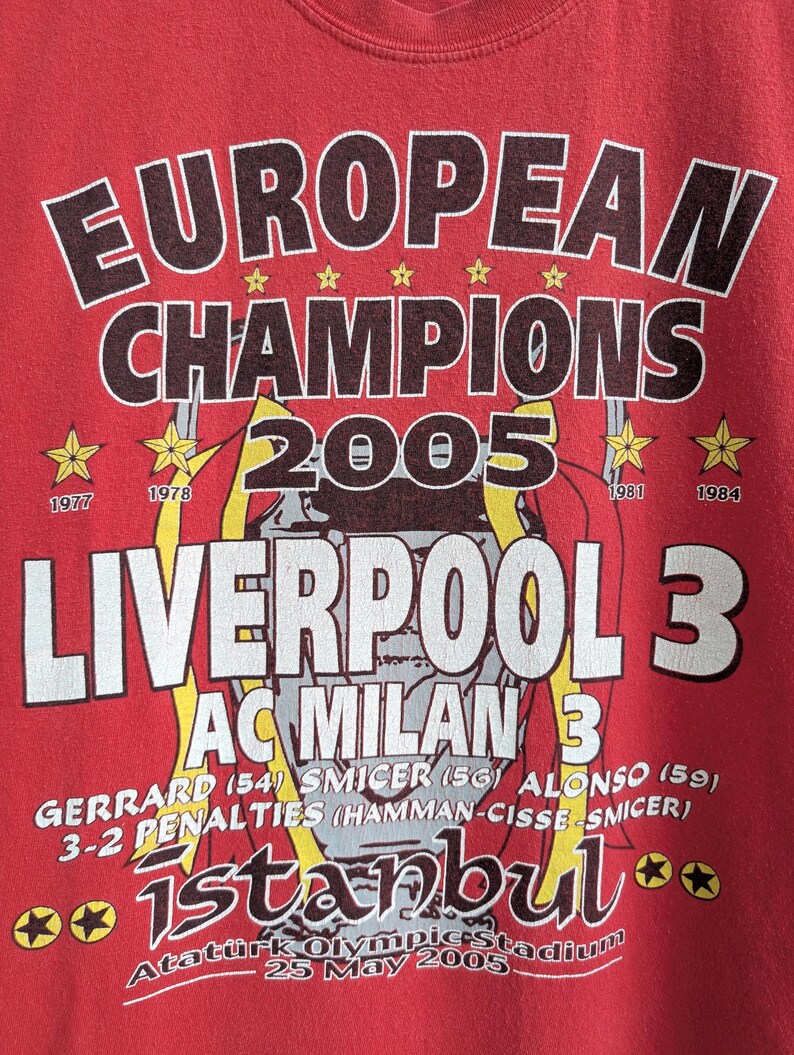 May include: Red t-shirt with a graphic commemorating Liverpool's 2005 Champions League victory. The design includes the words "EUROPEAN CHAMPIONS 2005", team names, scores, and the location and date of the match.