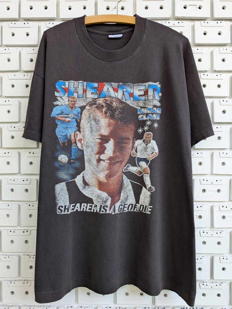 May include: Dark gray t-shirt featuring a graphic of a soccer player with the name "SHEARER" in blue and red. The text "SHEARER IS A GEORDIE" is also visible. The shirt is hanging on a wooden hanger.
