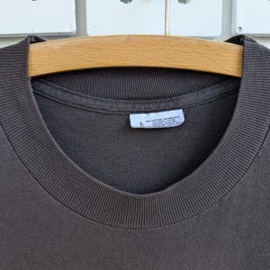 May include: Close-up of a dark brown t-shirt with a ribbed crew neck. The shirt is hanging on a wooden hanger. A white tag with text is visible near the neckline. The shirt is made of a soft, comfortable material.