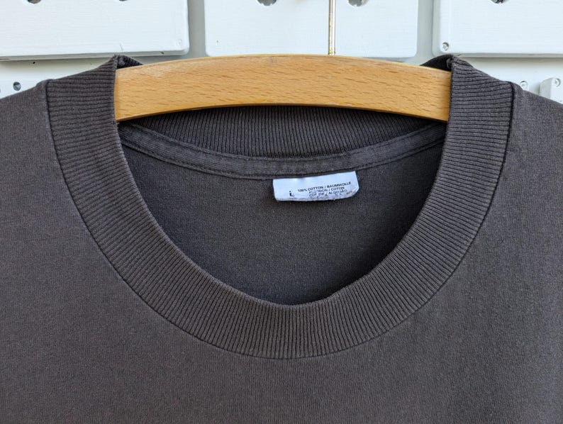 May include: Close-up of a dark brown t-shirt with a ribbed crew neck. The shirt is hanging on a wooden hanger. A white tag with text is visible near the neckline. The shirt is made of a soft, comfortable material.