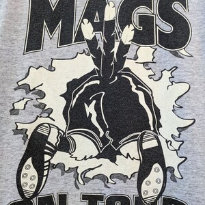 Puede incluir: Una camiseta gris con un gr&aacute;fico en blanco y negro de un personaje de dibujos animados que lleva un tocado con plumas. El texto "SUPER MAGS ON TOUR" est&aacute; impreso en la camiseta.