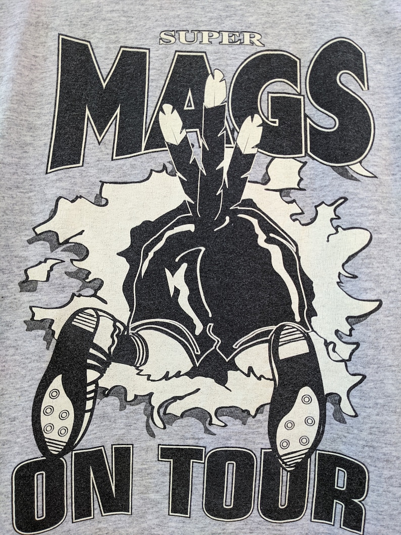 Puede incluir: Una camiseta gris con un gr&aacute;fico en blanco y negro de un personaje de dibujos animados que lleva un tocado con plumas. El texto "SUPER MAGS ON TOUR" est&aacute; impreso en la camiseta.