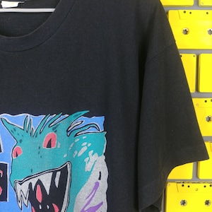 May include: Black t-shirt with a graphic print of a green monster with red eyes and sharp teeth. The monster is surrounded by blue, purple, and gray shapes.