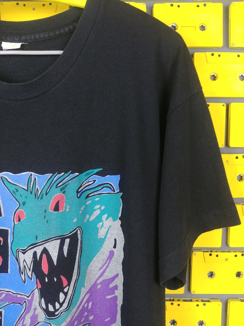 May include: Black t-shirt with a graphic print of a green monster with red eyes and sharp teeth. The monster is surrounded by blue, purple, and gray shapes.