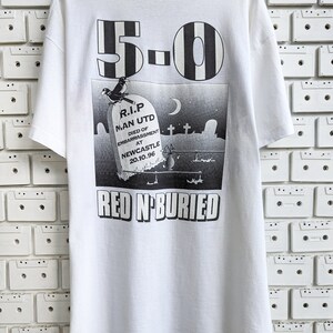 Pu&ograve; includere: Maglietta bianca con una stampa grafica in bianco e nero di una lapide con il testo "R.I.P. MAN UTD DIED OF EMBARRASSMENT AT NEWCASTLE 20.10.96" e il numero "5-0" sopra la lapide. Il testo "RED N-BURIED" &egrave; stampato sotto la lapide.