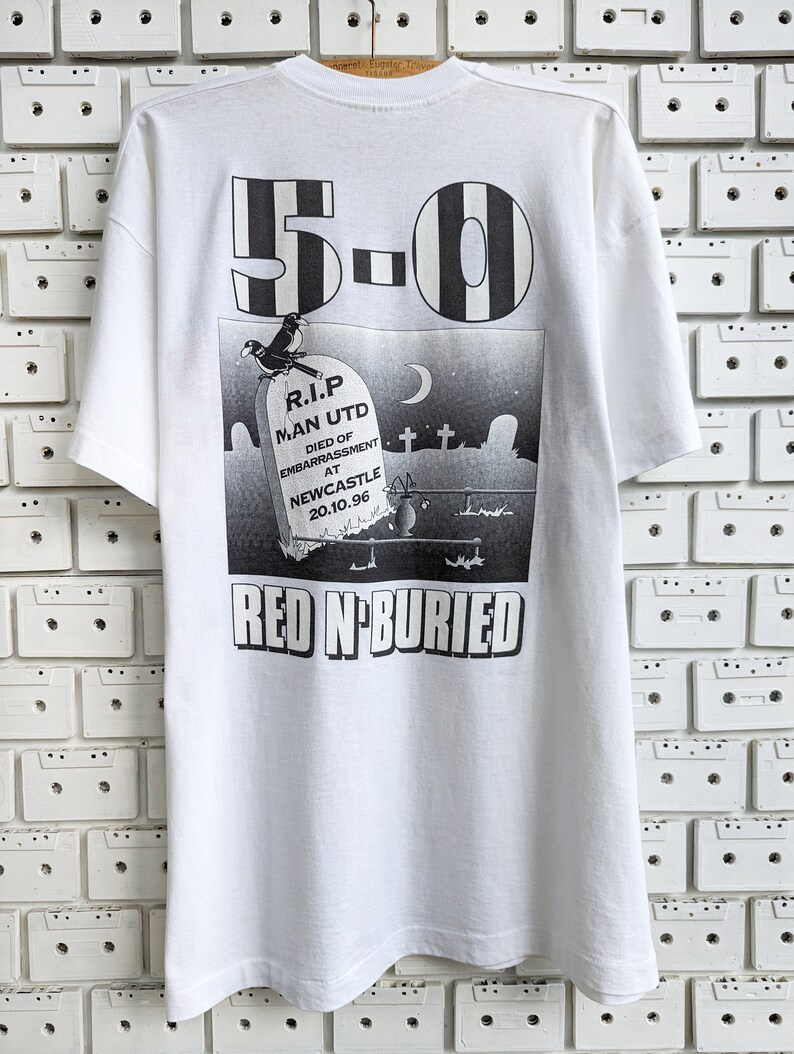Pu&ograve; includere: Maglietta bianca con una stampa grafica in bianco e nero di una lapide con il testo "R.I.P. MAN UTD DIED OF EMBARRASSMENT AT NEWCASTLE 20.10.96" e il numero "5-0" sopra la lapide. Il testo "RED N-BURIED" &egrave; stampato sotto la lapide.