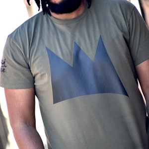 May include: Olive green t-shirt featuring a large, dark blue crown design on the front. The shirt has short sleeves and a crew neck. A small graphic is visible on the left sleeve. The shirt is made of a soft, comfortable material.