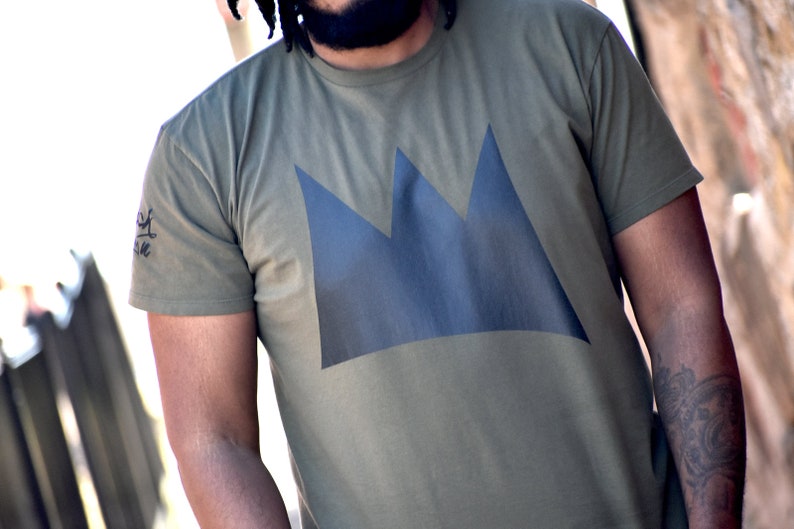 May include: Olive green t-shirt featuring a large, dark blue crown design on the front. The shirt has short sleeves and a crew neck. A small graphic is visible on the left sleeve. The shirt is made of a soft, comfortable material.