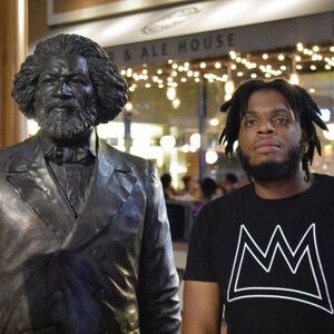 Puede incluir: Una estatua de bronce de un hombre con traje y corbata est&aacute; junto a una persona que lleva una camiseta negra con un dise&ntilde;o de corona blanca. El fondo incluye un letrero que dice "... & Ale House" y luces de cadena.