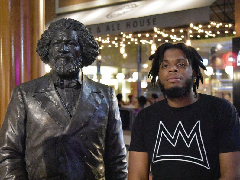 Puede incluir: Una estatua de bronce de un hombre con traje y corbata est&aacute; junto a una persona que lleva una camiseta negra con un dise&ntilde;o de corona blanca. El fondo incluye un letrero que dice "... & Ale House" y luces de cadena.