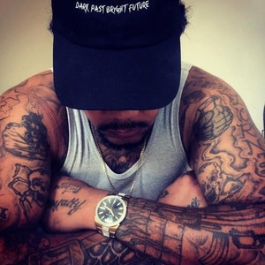 May include: A person wearing a black baseball cap with the text "DARK PAST BRIGHT FUTURE" in white. The person is wearing a gray tank top and a gold watch with a black face. The person has tattoos on their arms and chest.