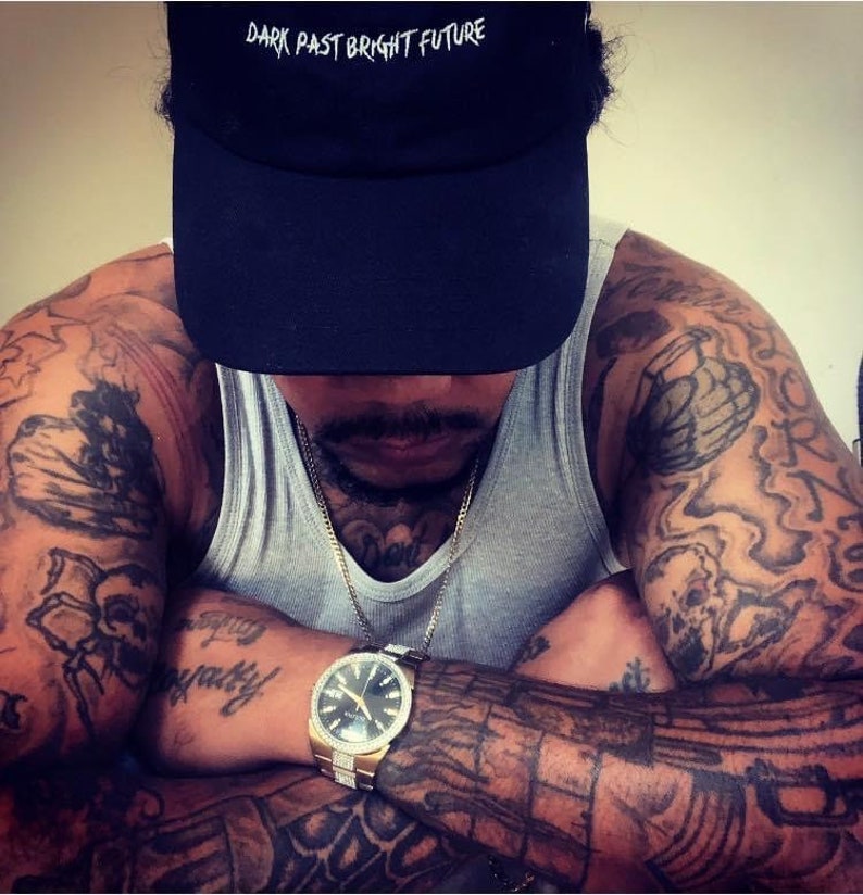 May include: A person wearing a black baseball cap with the text "DARK PAST BRIGHT FUTURE" in white. The person is wearing a gray tank top and a gold watch with a black face. The person has tattoos on their arms and chest.