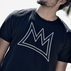 Puede incluir: Camiseta negra con un dise&ntilde;o de corona blanca. El dise&ntilde;o de la corona es una forma geom&eacute;trica simple. La camiseta tiene cuello redondo y mangas cortas, adecuada para el uso diario.