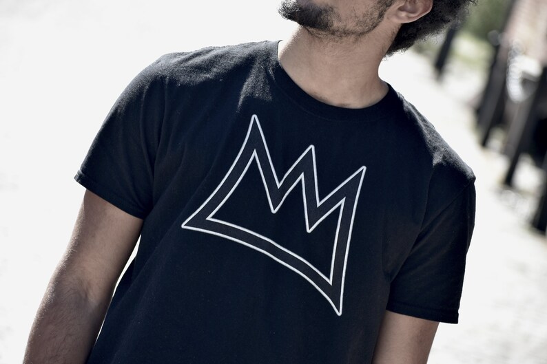 Puede incluir: Camiseta negra con un dise&ntilde;o de corona blanca. El dise&ntilde;o de la corona es una forma geom&eacute;trica simple. La camiseta tiene cuello redondo y mangas cortas, adecuada para el uso diario.