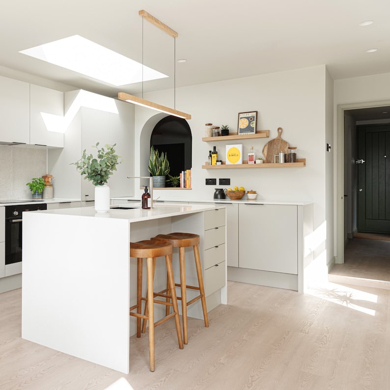 May include: A white kitchen island with a white worktop and two wooden bar stools. The island has a built-in drawer and is situated in a modern kitchen with white cabinets and a skylight.