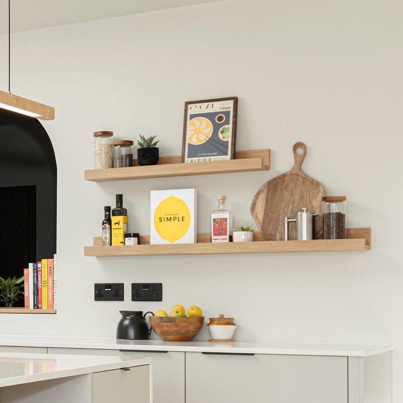 May include: Two wooden shelves mounted on a white wall with various items on them, including a cookbook titled "Simple", a framed print, jars of spices, a chopping board, and bottles of oil and vinegar.