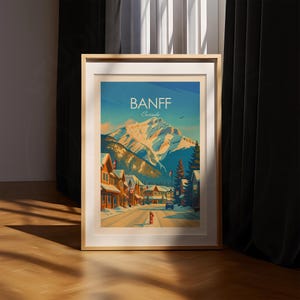Puede incluir: Un p&oacute;ster de viaje enmarcado que muestra una calle nevada en Banff, Canad&aacute;. La ilustraci&oacute;n representa un gran pico monta&ntilde;oso cubierto de nieve bajo un cielo azul, con edificios de madera, &aacute;rboles de hoja perenne y una boca de incendio roja.