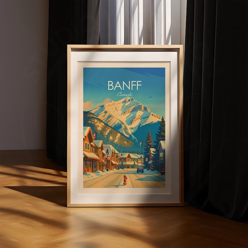 Puede incluir: Un p&oacute;ster de viaje enmarcado que muestra una calle nevada en Banff, Canad&aacute;. La ilustraci&oacute;n representa un gran pico monta&ntilde;oso cubierto de nieve bajo un cielo azul, con edificios de madera, &aacute;rboles de hoja perenne y una boca de incendio roja.