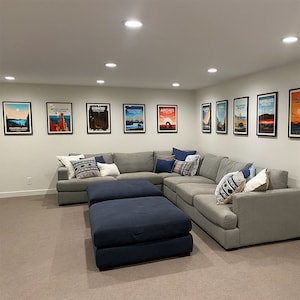 May include: A modern living room featuring a large gray sectional sofa with decorative pillows and two navy blue ottomans. Framed travel posters of national parks hang on the white walls, creating a cozy and inviting space.