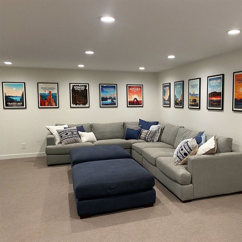 May include: A modern living room featuring a large gray sectional sofa with decorative pillows and two navy blue ottomans. Framed travel posters of national parks hang on the white walls, creating a cozy and inviting space.