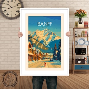 Puede incluir: Una impresi&oacute;n art&iacute;stica enmarcada de Banff, Canad&aacute;, que presenta una calle cubierta de nieve bordeada de edificios y una majestuosa cordillera en el fondo. La obra de arte tiene un estilo de cartel de viaje vintage con un cielo azul y la palabra "Banff" en la parte superior.